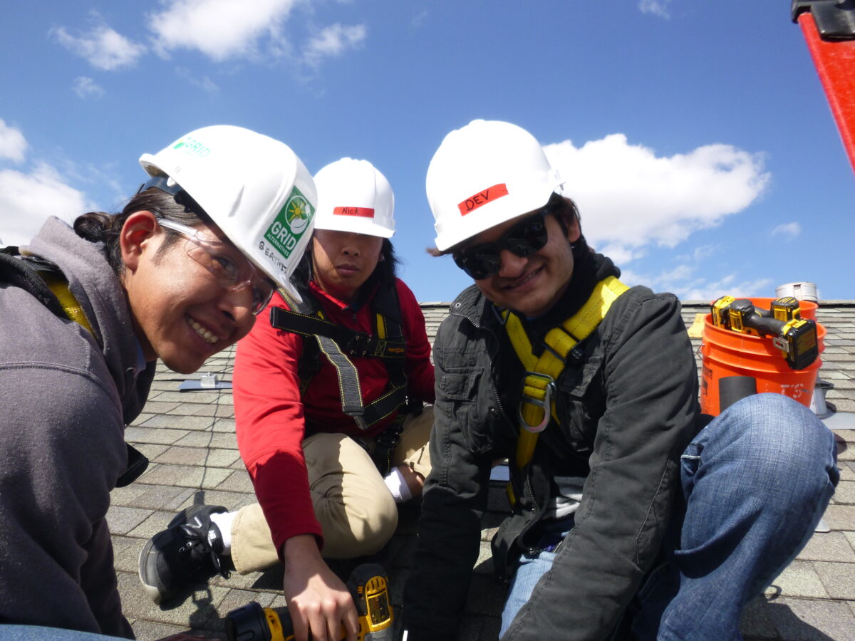 Three workers in hard hats and safety gear examine roofing materials on a sunny residential roof