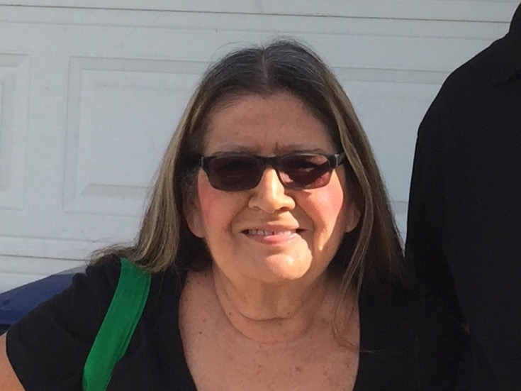 Woman wearing sunglasses and dark jacket with green shoulder strap, smiling outdoors