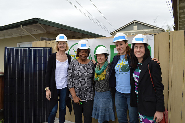 Five women wearing white hard hats stand together in front of solar panels at a construction site