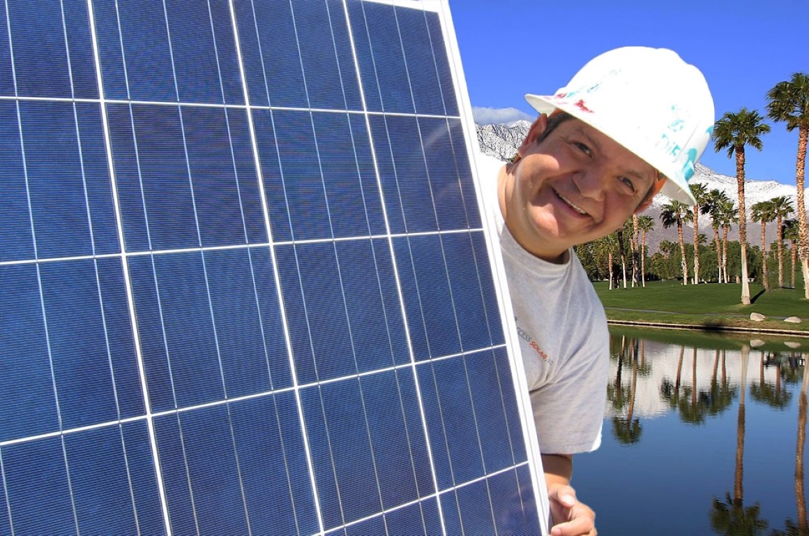 Worker in white hard hat smiling behind large solar panel with palm trees and water in background
