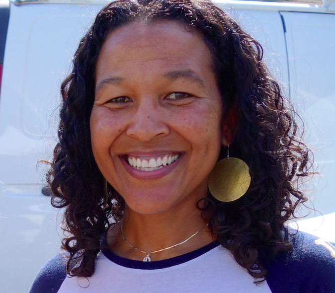 Smiling woman with curly dark hair and gold hoop earrings wearing a blue and white shirt