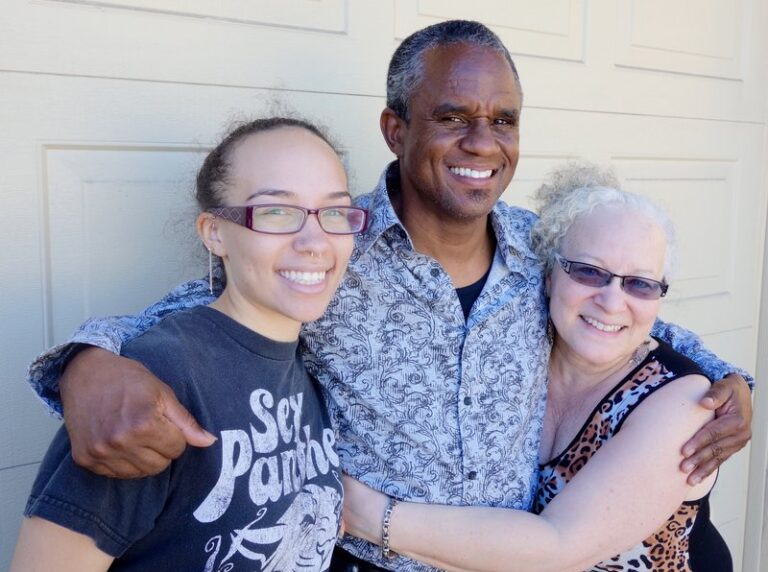 Three smiling people posing together in front of a white garage door, appearing happy and excited