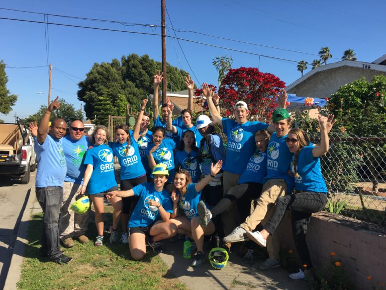 Large volunteer group in blue GRID shirts celebrating outdoors, arms raised in joy, with blooming red flowers visible in background