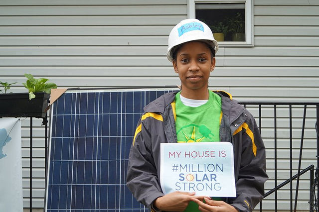 Teen in hard hat holding sign "My House Is #MillionSolarStrong" in front of home solar panel
