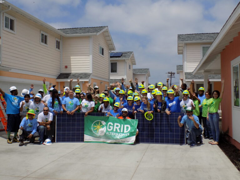 Large group of volunteers in hard hats and GRID Alternatives shirts holding solar panels in front of new homes