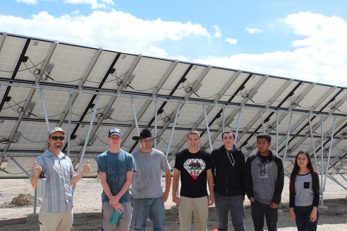 Seven young people standing in front of large solar panel array under blue sky
