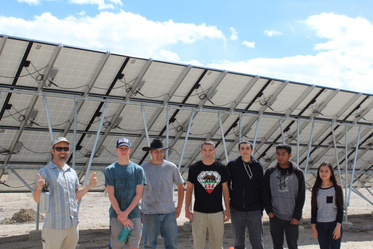 Seven young people standing in front of large solar panel array under blue sky