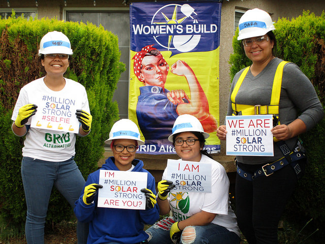 Five people in hard hats holding signs promoting "#MillionSolarStrong" in front of a Women's Build banner with Rosie the Riveter imagery