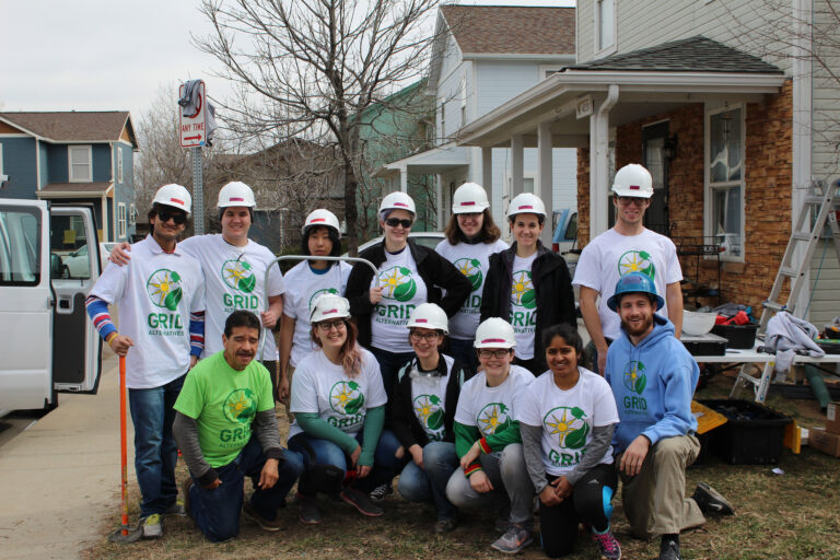 Team wearing GRID Alternative hard hats and matching t-shirts posing together at a residential construction site