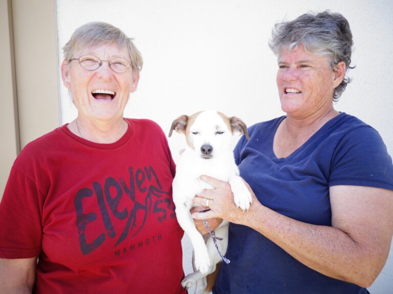 Two women and a white dog with brown markings smile together indoors