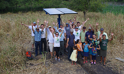 Community group gathered outdoors around solar panel installation with raised hands in celebration
