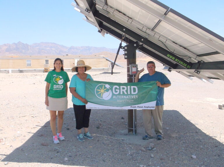 Three people holding GRID Alternatives Inland Empire banner at solar installation site in desert
