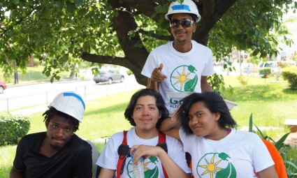 Four young people wearing matching white t-shirts and blue hard hats pose together outdoors under a tree