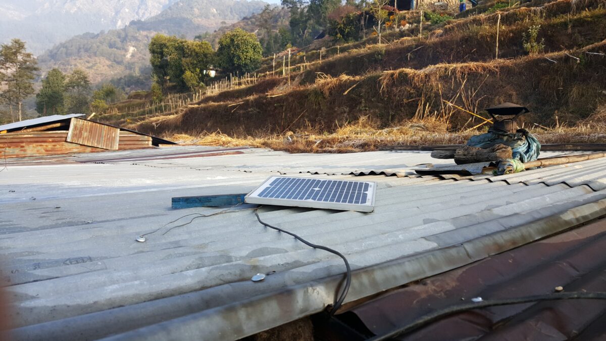 Small solar panel on corrugated metal roof in mountainous rural Nepal with terraced fields and wooden structures in background