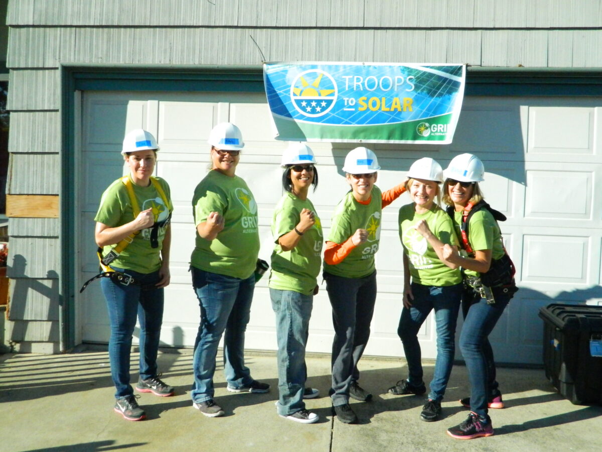 Six women in green shirts and hard hats pose in front of a "Troops to Solar" banner on a building