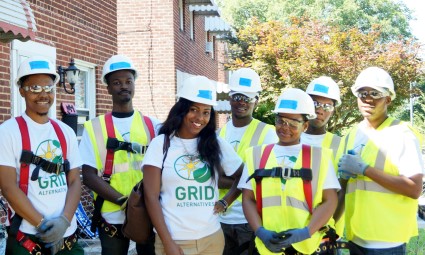 Six construction workers wearing hard hats and safety vests pose together outdoors, one wearing a GRID t-shirt