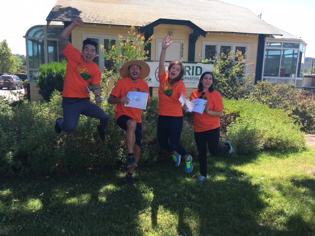 Four people in orange shirts jumping in front of a house holding papers, with "RID" sign visible