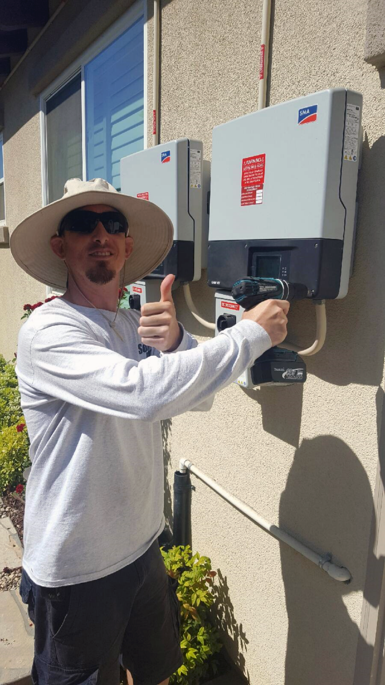 Man in cowboy hat giving thumbs up beside two white solar inverter units mounted on building wall