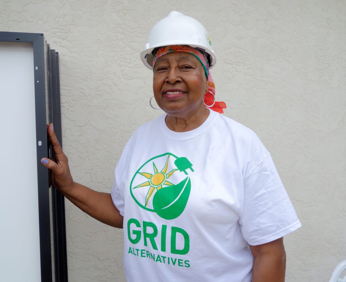 Woman wearing GRID Alternatives shirt and hard hat, holding solar panel frame against white wall