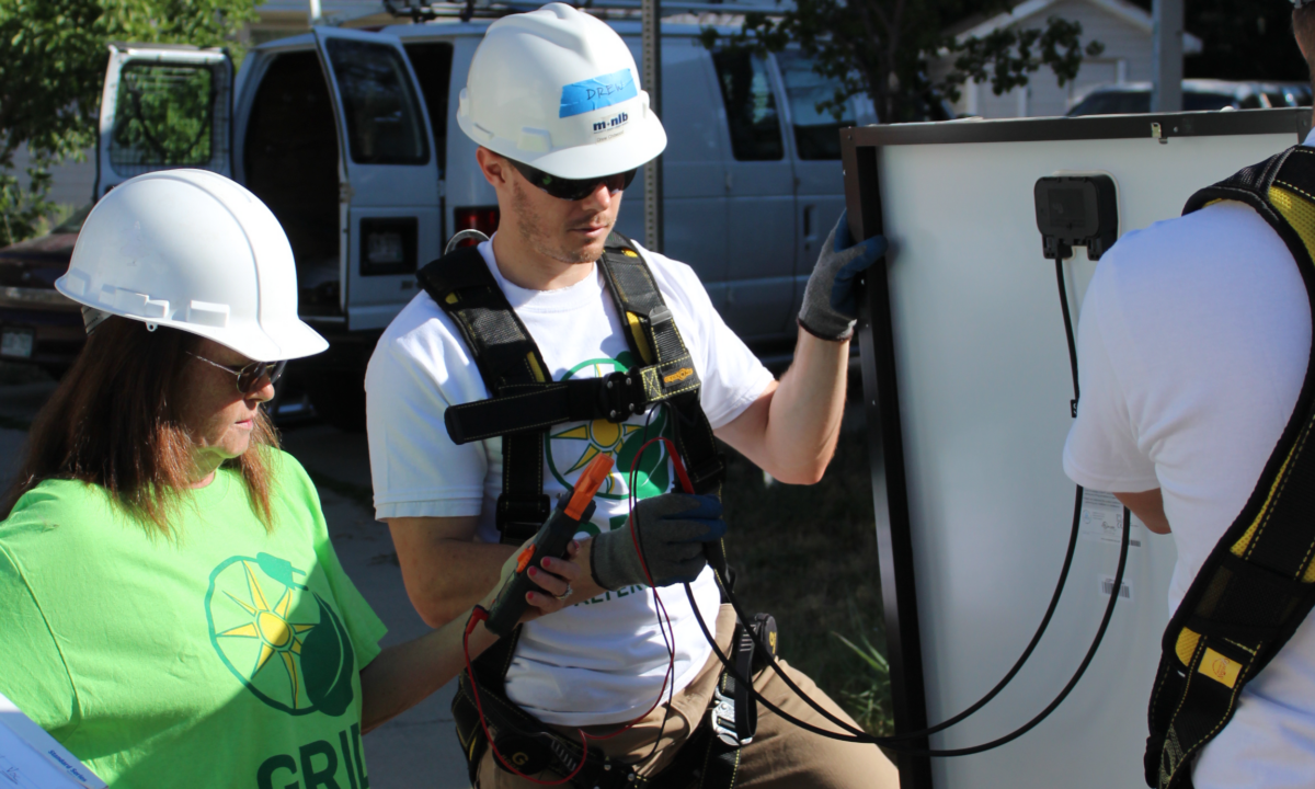 Two workers in hard hats and safety harnesses installing electrical equipment on a white panel outdoors