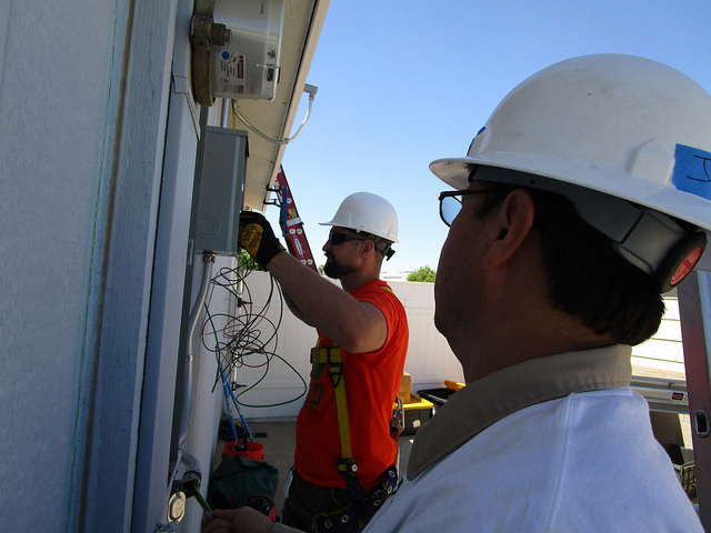 Two construction workers in hard hats installing electrical equipment on a building exterior during daytime