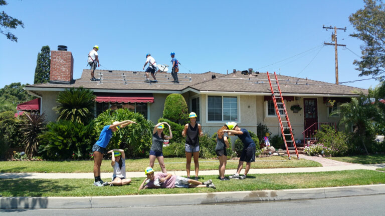 Community volunteers in hard hats working on roof repairs and exterior renovation of a residential house