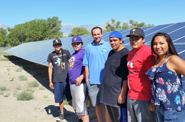 Six people standing in front of a large solar panel array with mountains and trees in the background