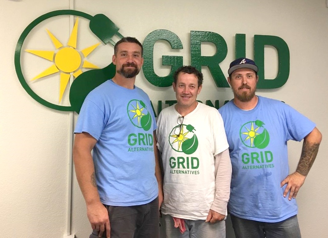 Three men wearing GRID Alternatives branded t-shirts stand in front of GRID Alternatives logo on white wall