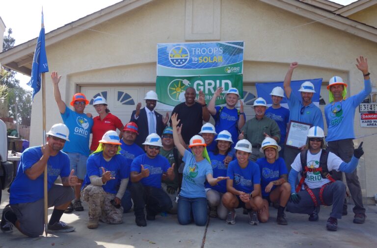 Group of volunteers in blue GRID Alternatives shirts and hard hats posing in front of house with "Troops to Solar" banner