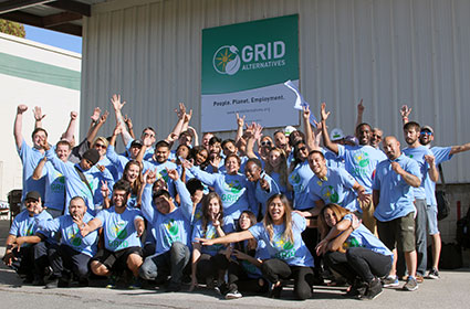 Large group of SolarCorps Fellows in blue shirts raising hands in celebration under GRID Alternatives banner
