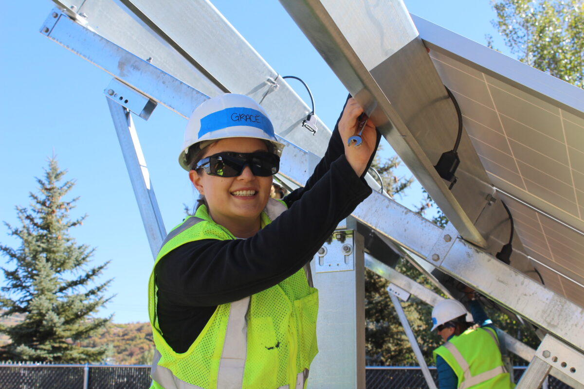 Young worker in safety gear and hard hat smiling while installing solar panels outdoors