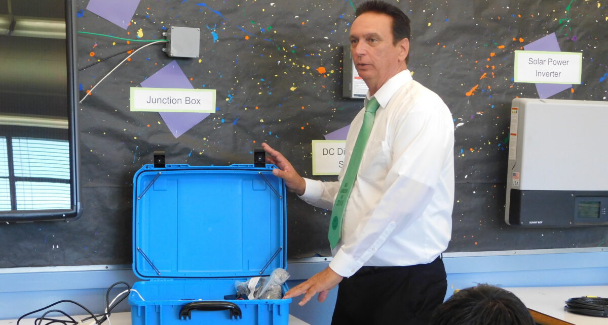 Man presenting solar power system components with blue case in front of diagram showing junction box, DC disconnect, and solar power inverter
