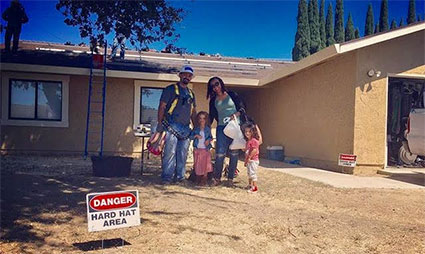 Family standing in front of single-story building with "Danger Hard Hat Area" sign visible in yard
