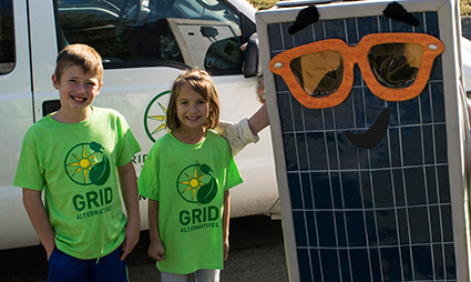 Two children in green GRID shirts stand next to a solar panel with large orange sunglasses on it