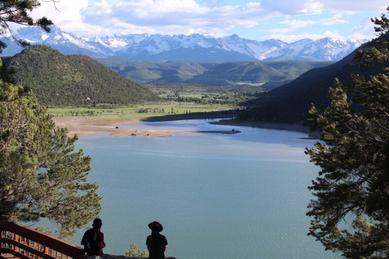 Two hikers overlook a turquoise alpine lake surrounded by forested mountains and snowy peaks