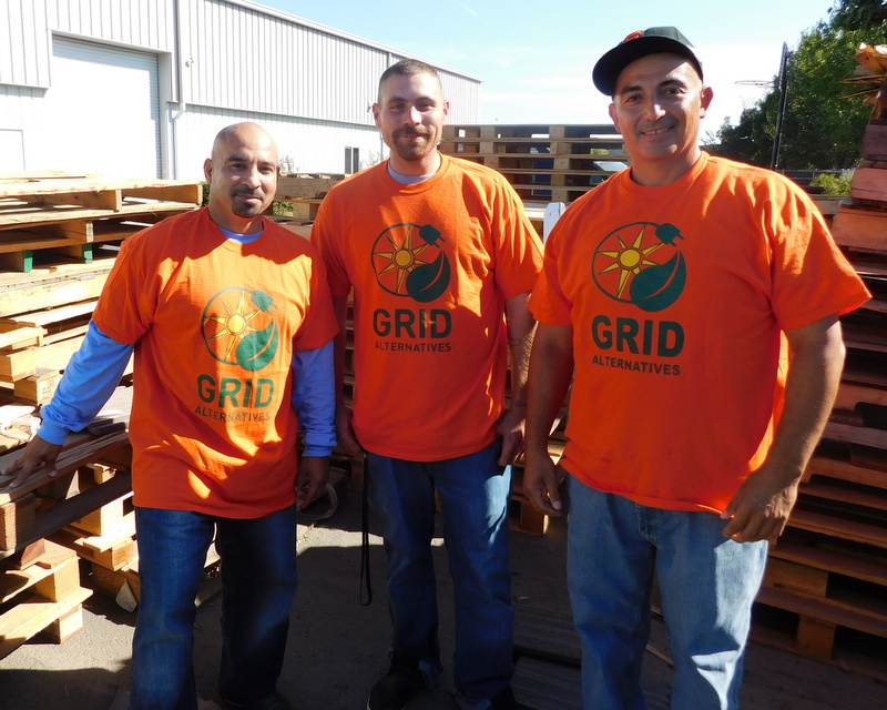Three men wearing orange GRID Alternatives t-shirts stand together at a work site with lumber in the background