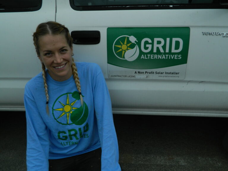 Woman in GRID Alternatives shirt smiling in front of white van with company logo and "A Non Profit Solar Installer" text