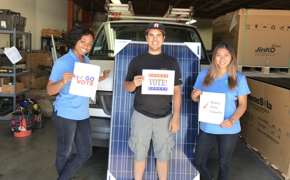 Three people hold voting signs in front of a solar panel on a truck bed in a warehouse setting