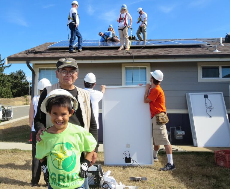 Community volunteers install solar panels on a residential roof while a child in a green "Grill Alternatives" shirt smiles at the camera