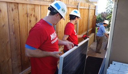 Volunteers in red shirts and hard hats install solar panels on a home exterior