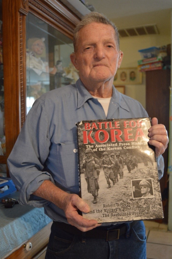 Elderly man holding "Battle for Korea" book by Associated Press in home interior
