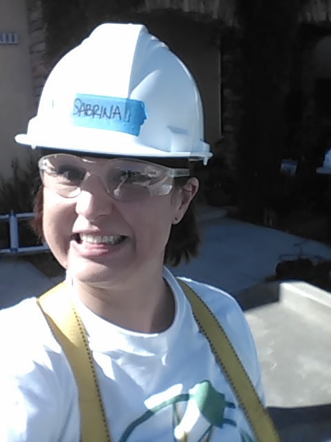 Woman wearing white hard hat with "Sapphia" text, safety glasses, and work vest, smiling at camera