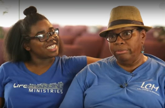 Two women in blue ministry shirts smiling together, one wearing glasses and a tan hat