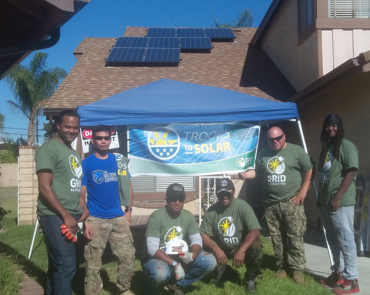 Group of volunteers in green shirts standing under blue tent with "Troops to Solar" banner, solar panels visible on house roof behind them