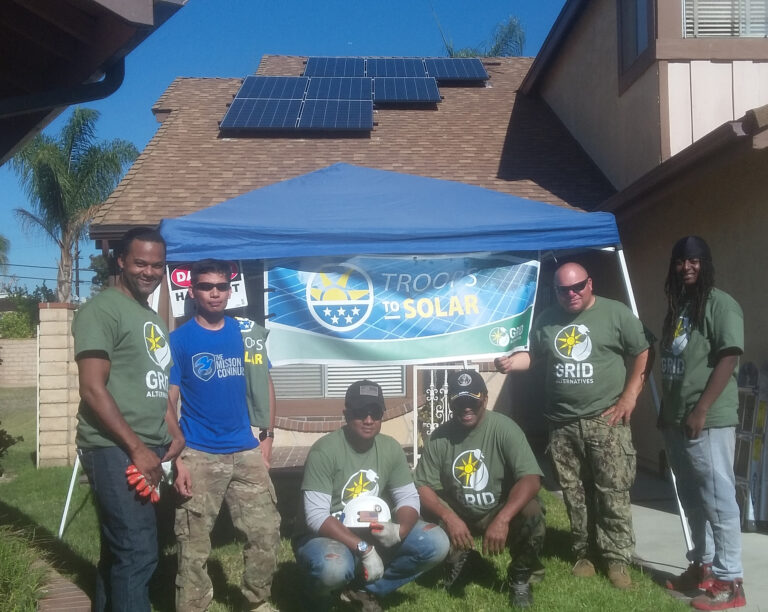 Group of volunteers in green shirts standing under blue tent with "Troops to Solar" banner, solar panels visible on house roof behind them