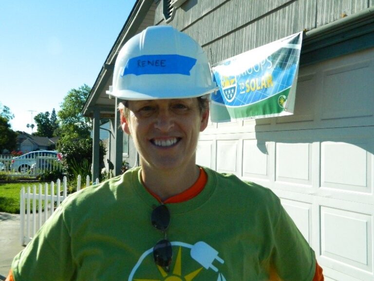 Man in green shirt wearing white hard hat with "RENEE" text, standing in front of solar panel flag