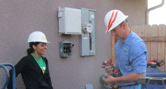 Two construction workers in hard hats discussing plans near an electrical meter box on a building wall