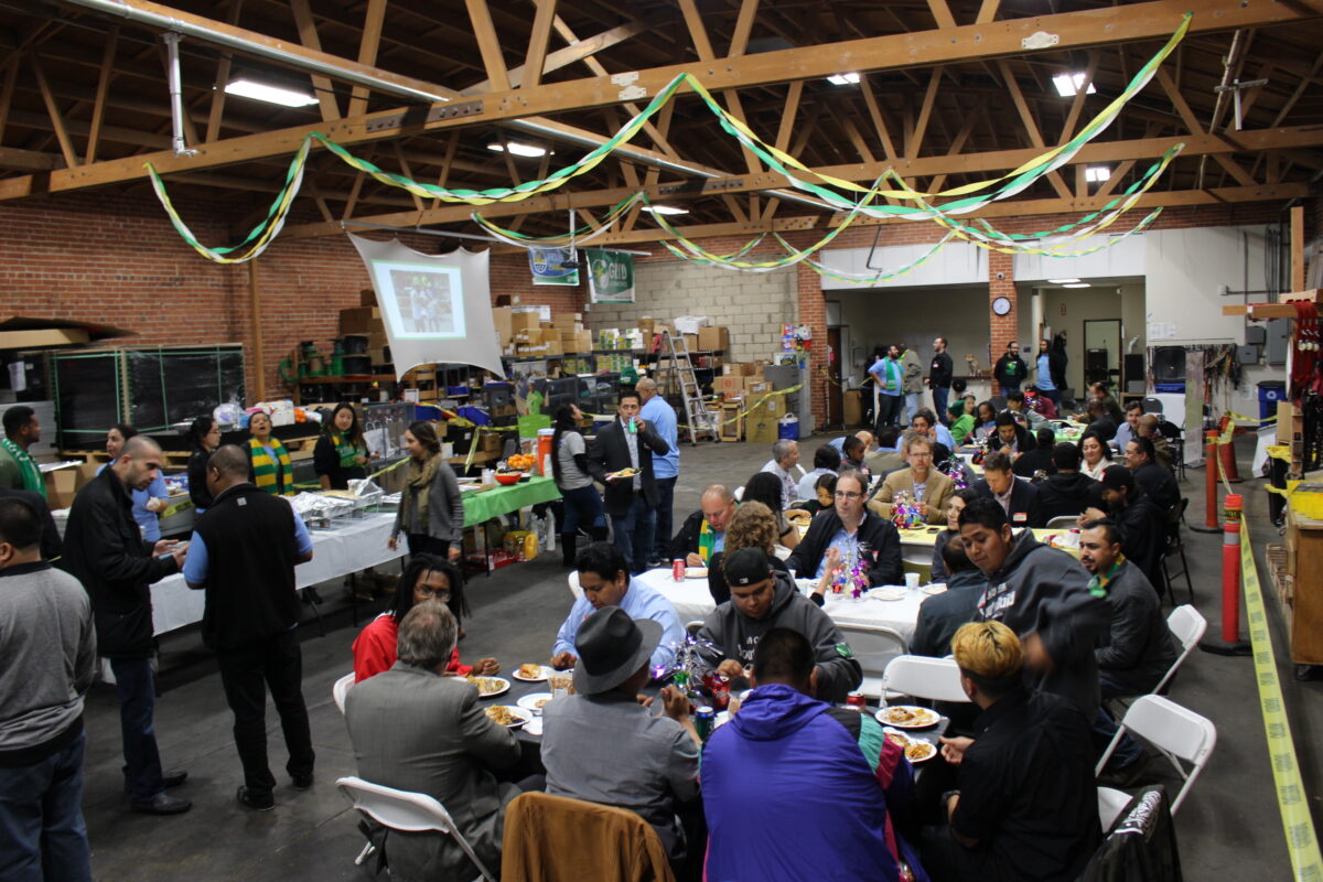 Community gathering in a gymnasium with people seated at tables eating, green and gold decorations overhead, food service stations in background