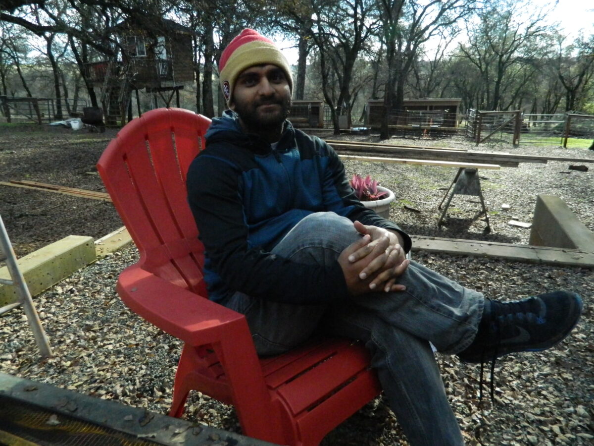 Man with beard wearing red Adirondack chair at ranch with trees in background