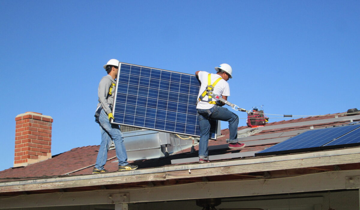 Two workers in hard hats installing a solar panel on a residential roof under clear blue sky
